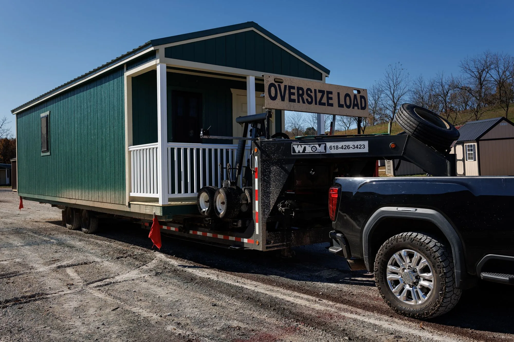 A green cabin with a white porch loaded on a delivery trailer behind a pickup truck