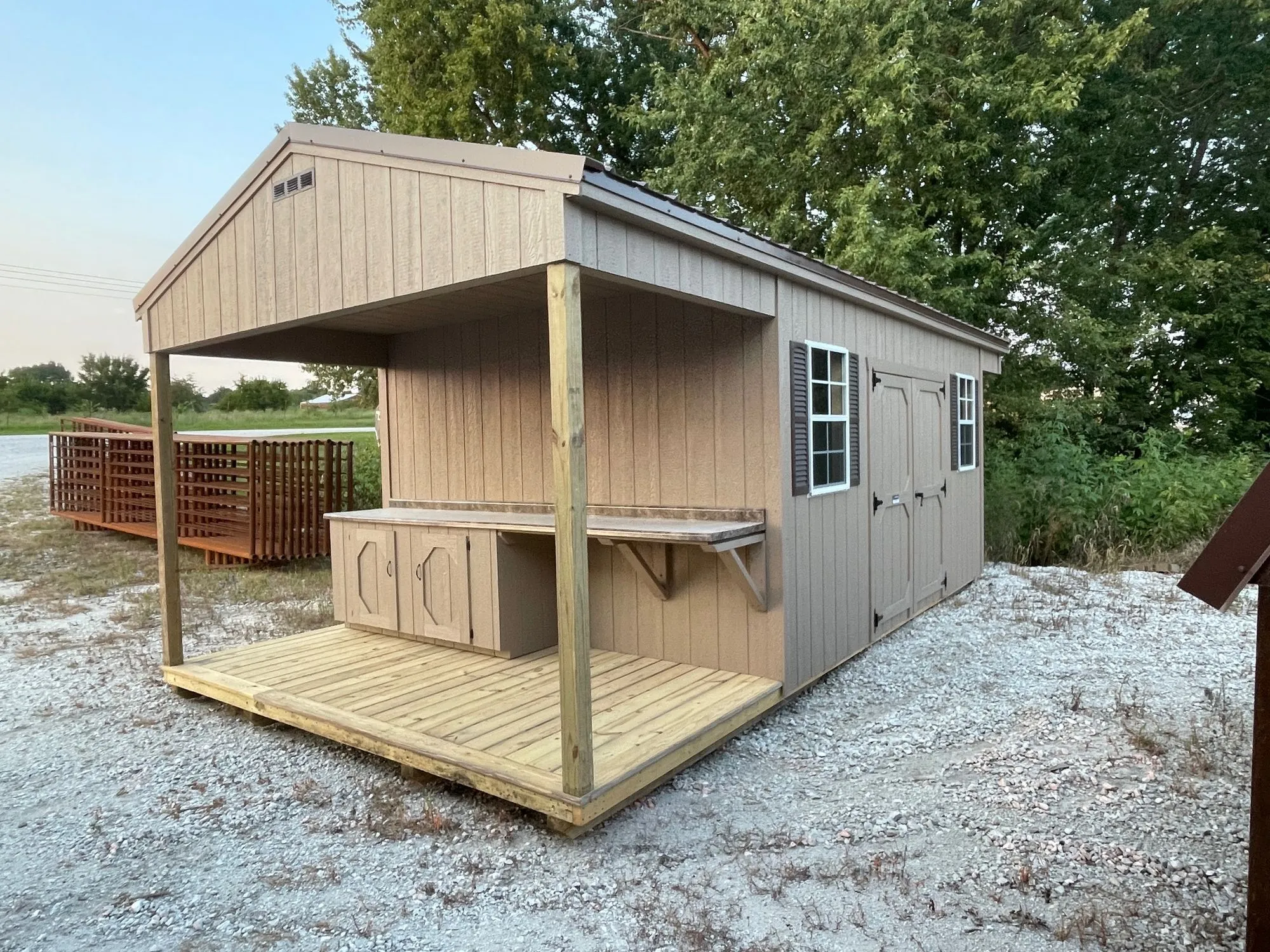 A tan gable shed with white-framed windows, a covered wood porch and a potting bench with cabinets under it